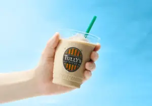 Hand holding a plastic iced coffee cup with Tullys Coffee logo against a bright blue background