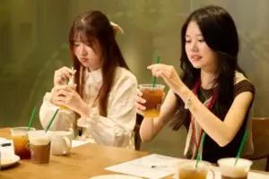 Two women sit at a cafe table each holding a cup with a green straw and adjusting their drinks