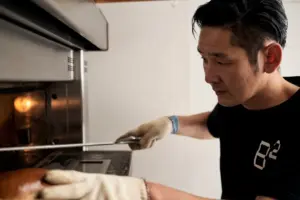 5941b4a1b2f010defdc0a4e68a7b1f63 | コーヒーステーション Man wearing gloves uses a long tool to place or remove a tray from a stainless steel oven
