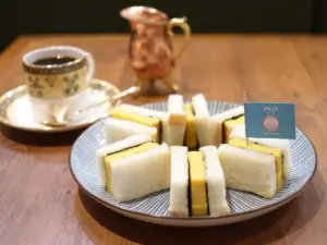 Plate of mini white bread egg sandwiches arranged on a striped plate with a blue flag label tea cup and copper pitcher in the background for tea time snacking