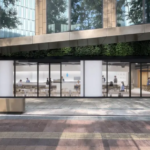 Modern glass front cafe with interior seating and customers set in a urban plaza with a bench and greenery outside