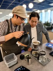 Barista in apron and cap pours hot water over a pour over coffee setup as a man in a suit watches nearby