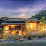 Traditional wooden Japanese house lit warmly at dusk with a landscaped garden and hills in the background