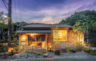 Traditional wooden Japanese house lit warmly at dusk with a landscaped garden and hills in the background