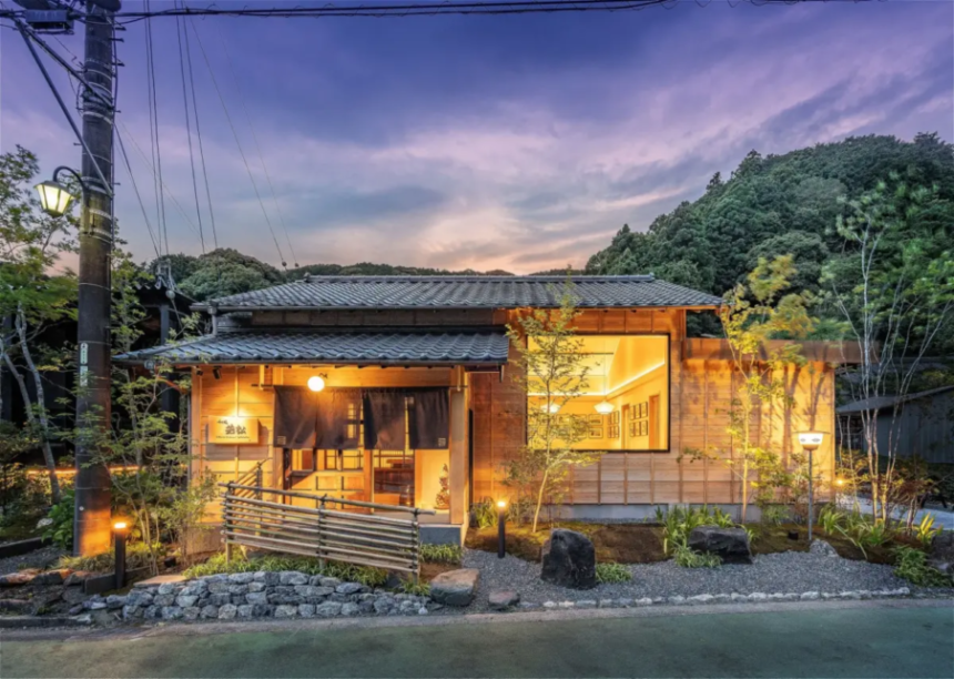 Traditional wooden Japanese house lit warmly at dusk with a landscaped garden and hills in the background