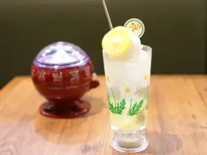 Tall glass of lemonade with ice and a lemon wheel featuring a daisy pattern on the glass placed on a wooden table with a red ceramic pot blurred in the background