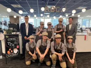 Group of cafe staff in checkered shirts and beige caps posing with two managers in suits at a cafe counter under an ABC cafe sign