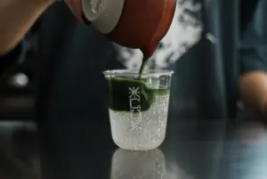 Close up of a hand pouring a green liquid from a shaker into a clear cup with ice and fizzy bubbles