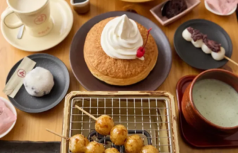 A spread of Japanese desserts and snacks on a wooden table: a pancake with whipped cream, a fruit platter, mochi and sweets, skewer-grilled fried dough balls, and bowls of matcha and milk tea.