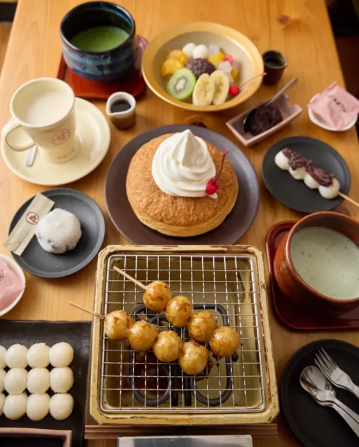 A spread of Japanese desserts and snacks on a wooden table a pancake with whipped cream a fruit platter mochi and sweets skewer grilled fried dough balls and bowls of matcha and milk tea