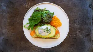 Open faced toast topped with avocado and white fish surrounded by mixed greens and shredded carrot on a white plate