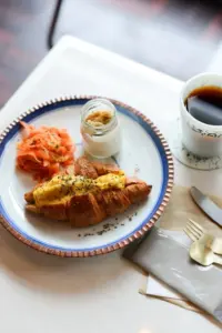 Brunch plate with a croissant topped with melted cheese and herbs smoked salmon on the side a small jar of sauce and a cup of coffee