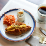 Plate with croissant topped with egg smoked salmon a jar of jam and a cup of coffee on a white table setting