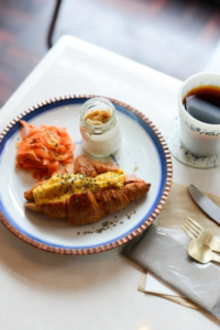 Plate with croissant topped with egg smoked salmon a jar of jam and a cup of coffee on a white table setting