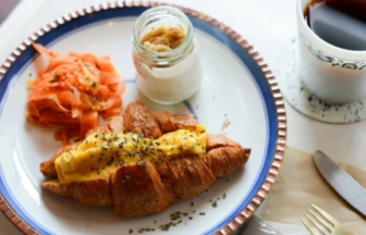 Plate with croissant topped with egg smoked salmon a jar of jam and a cup of coffee on a white table setting