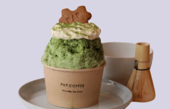 Green matcha shaved-ice dessert in a brown paper cup, topped with whipped cream and a cookie on top, served on a plate.