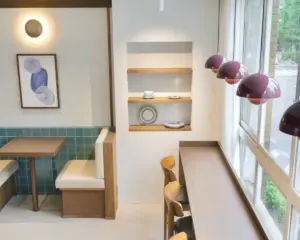 Modern cafe corner with teal tiled wall wooden tables and white cushioned seating a builtin shelf displaying dishes and a window counter with wooden stools under purple pendant lights