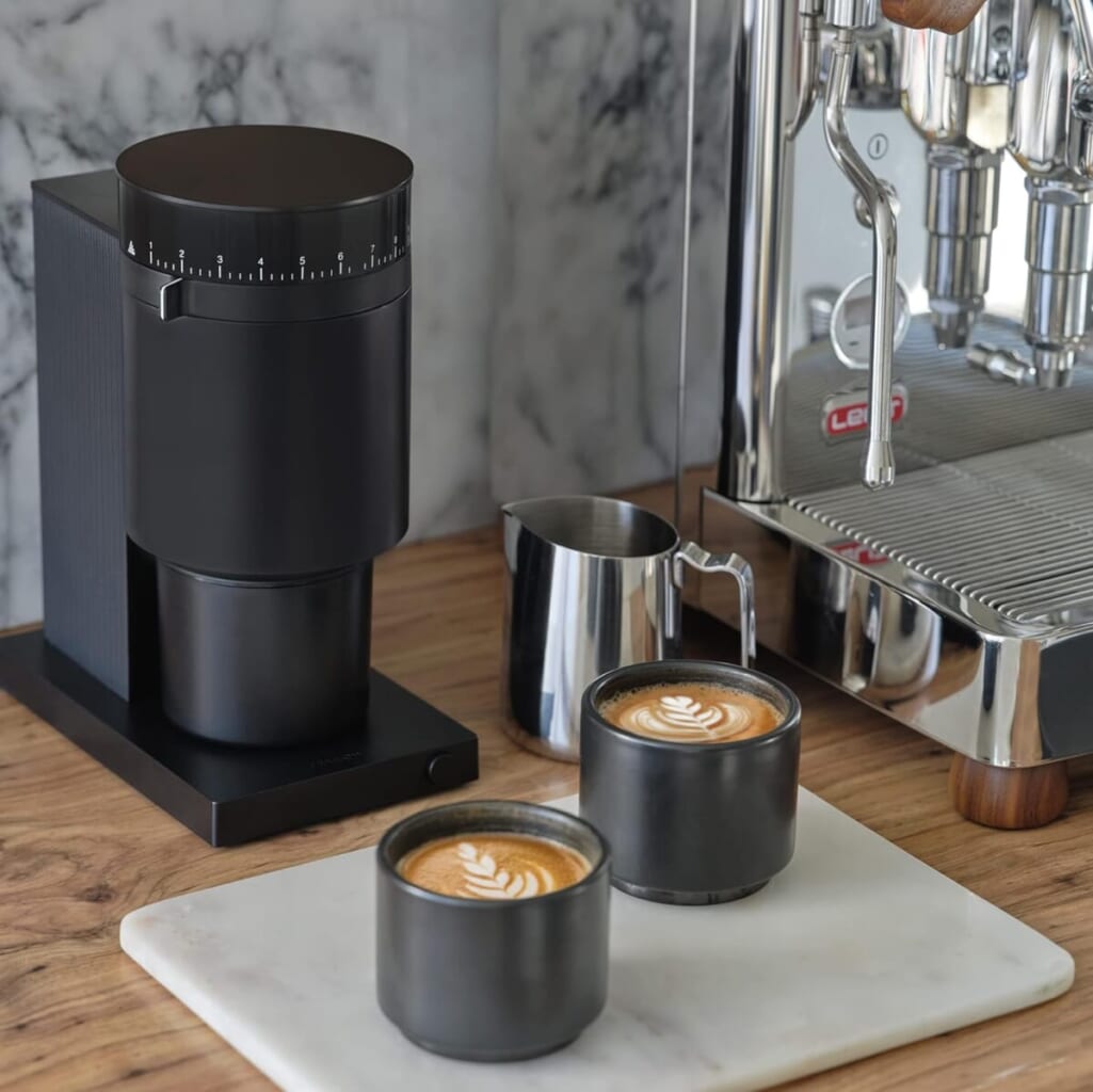 Two black cups with latte art sit on a white marble board next to a shiny espresso machine and a black coffee grinder on a wooden counter