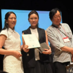 Three people stand on a stage center woman holds a certificate and a small award flanked by a smiling woman in white and a man with a badge Blue backdrop behind them