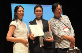 Three people stand on a stage; center woman holds a certificate and a small award, flanked by a smiling woman in white and a man with a badge. Blue backdrop behind them.