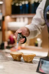 Chef uses a culinary torch to caramelize the tops of two small pastry tarts on a stainless counter