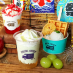 Bright dessert spread two milk based drinks in cups with whipped toppings fruit topping and waffles set on a wooden table with colorful festival banners and fireworks illustrations in the background plus green grapes nearby