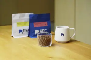 Glass cup filled with roasted coffee beans on a wooden table with white and blue coffee bags and a white mug in the background
