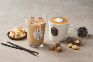 Iced coffee in a glass with ice next to a white mug of latte surrounded by popcorn chocolates vanilla pods and berries on a light surface