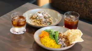 Two glasses of iced coffee and two bowls of food on a wooden table pasta with meat and grated cheese and a bowl of yellow rice with vegetables and naan