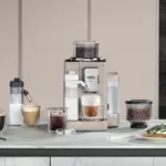 Espresso machine on a kitchen counter with several coffee drinks a grinder and a plate of toast on the right side