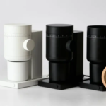 Three coffee siphon brewers in white and black on a white surface with a wooden lidstand to the right and a power cord visible behind them