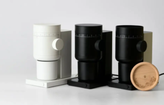 Three coffee siphon brewers in white and black on a white surface with a wooden lidstand to the right and a power cord visible behind them