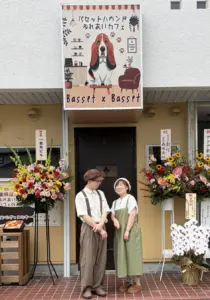 Two people in retro outfits stand outside a cafe entrance with a large Basset x Basset sign and colorful flower arrangements