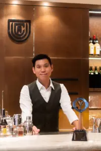 Bartender in a white shirt and dark vest stands behind a bar smiling with bottles and bar tools on the counter and a wall logo in the background