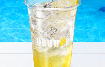Clear plastic cup of iced lemon drink with a lemon wedge ice and bubbles on a white surface beside a blue pool