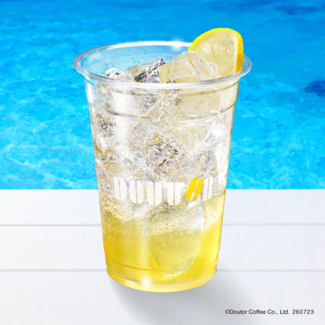 Clear plastic cup of iced lemon drink with a lemon wedge ice and bubbles on a white surface beside a blue pool
