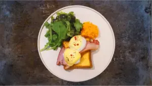 Plate of eggs Benedict with ham on toast two poached eggs with hollandaise alongside a green salad and a scoop of shredded carrot on a white plate