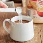 A hand pours powdered creamer into a white mug of coffee on a wooden table with a Coffee Mate canister in the background