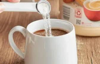 A hand pours powdered creamer into a white mug of coffee on a wooden table, with a Coffee Mate canister in the background.
