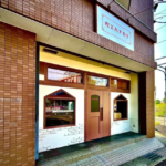 Cafe storefront with a pink door and diamond shaped windows set in a brick facade sign above in Japanese 2 windows flank the door under a white brick wall detail
