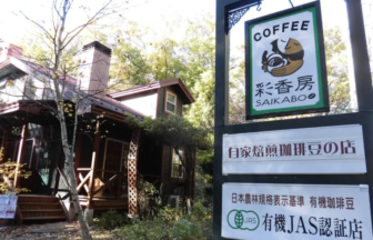 Exterior of a wooden cafe with a porch and trees next to a tall sign reading COFFEE SAIKABO