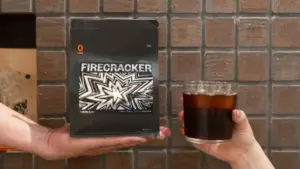 ac912b9f59958ac10bb84bc60ed0f94c | コーヒーステーション Two hands exchange a black Firecracker package for a glass of dark cola against a brick wall background