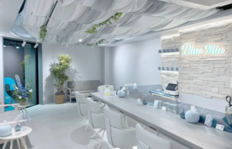 Bright modern salon or reception area with a long white counter white chairs plants and a glowing Blue Mio sign on a textured wall