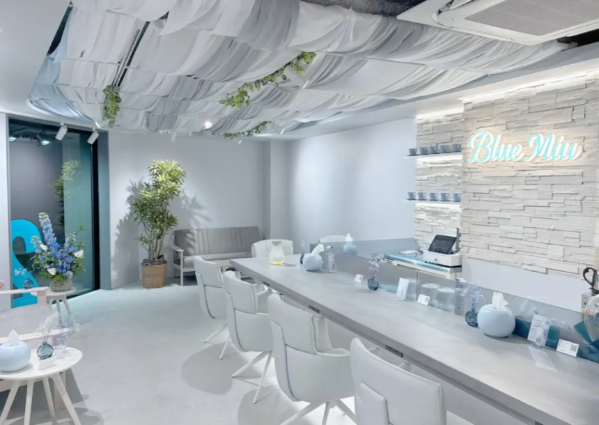 Bright modern salon or reception area with a long white counter white chairs plants and a glowing Blue Mio sign on a textured wall