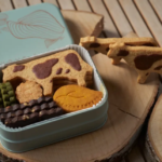 Open light blue tin filled with assorted cookies cow pattern biscuits green wafers chocolate bars on a wooden surface