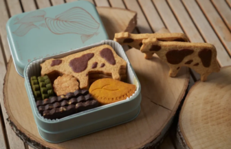 Open light-blue tin filled with assorted cookies (cow-pattern biscuits, green wafers, chocolate bars) on a wooden surface.