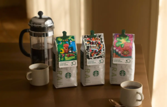 French press coffee maker on a wooden table with three colorful coffee bags and two cups nearby, sunlit scene.