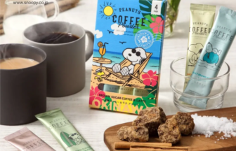 Peanuts Coffee Powder packaging with Snoopy artwork, two mugs of coffee, and sachets on a light table with a wooden board holding brown sugar chunks and a cinnamon spoon.