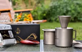 Outdoor camping coffee setup on a metal table: stainless steel pots, a pour-over dripper, and a foil-wrapped bag beside a black decorative box in a garden setting