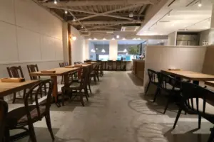 Interior of a minimalist cafe with wooden tables and chairs concrete floor and an open kitchen area in the back under warm pendant lighting